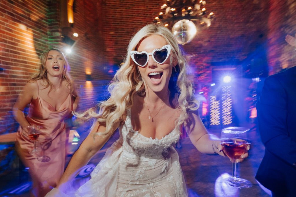 A joyful bride in heart-shaped sunglasses and a white lace dress dances at a lively indoor wedding reception, holding a drink, with another smiling guest in a pink dress behind her.