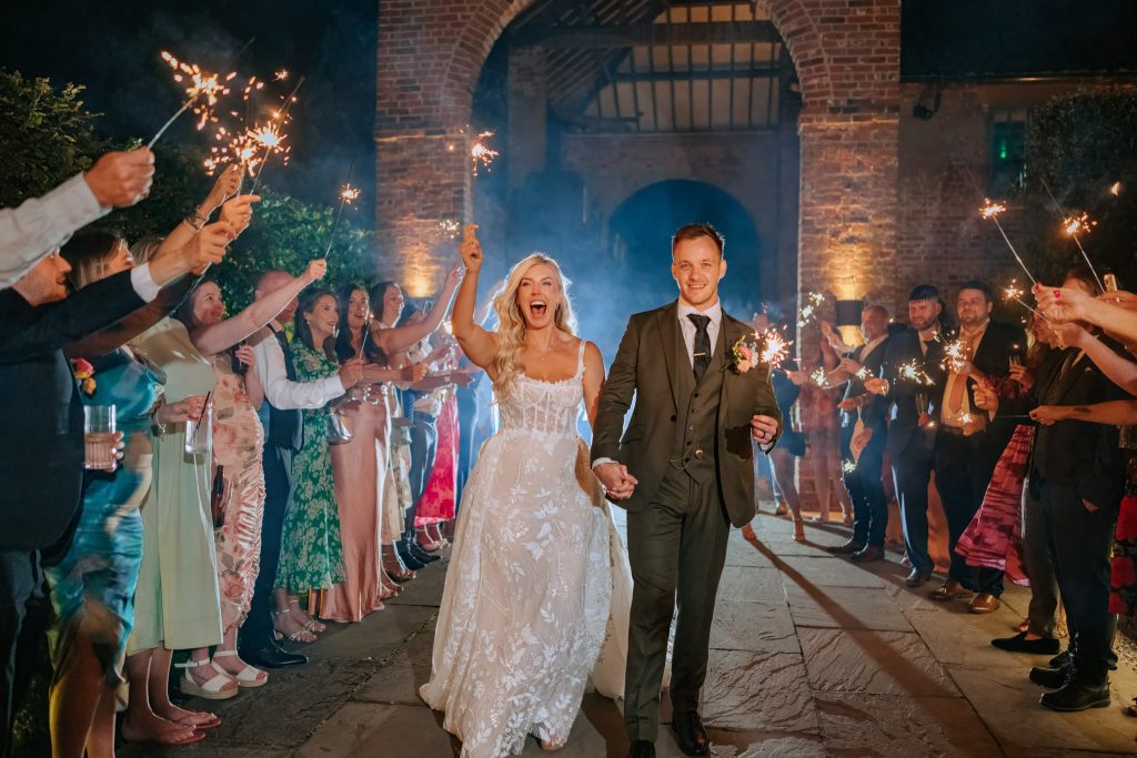 A joyful bride and groom walk hand-in-hand through a crowd of guests holding sparklers at night, celebrating their wedding. The couple is smiling, and the scene is lively and festive with warm lighting.
