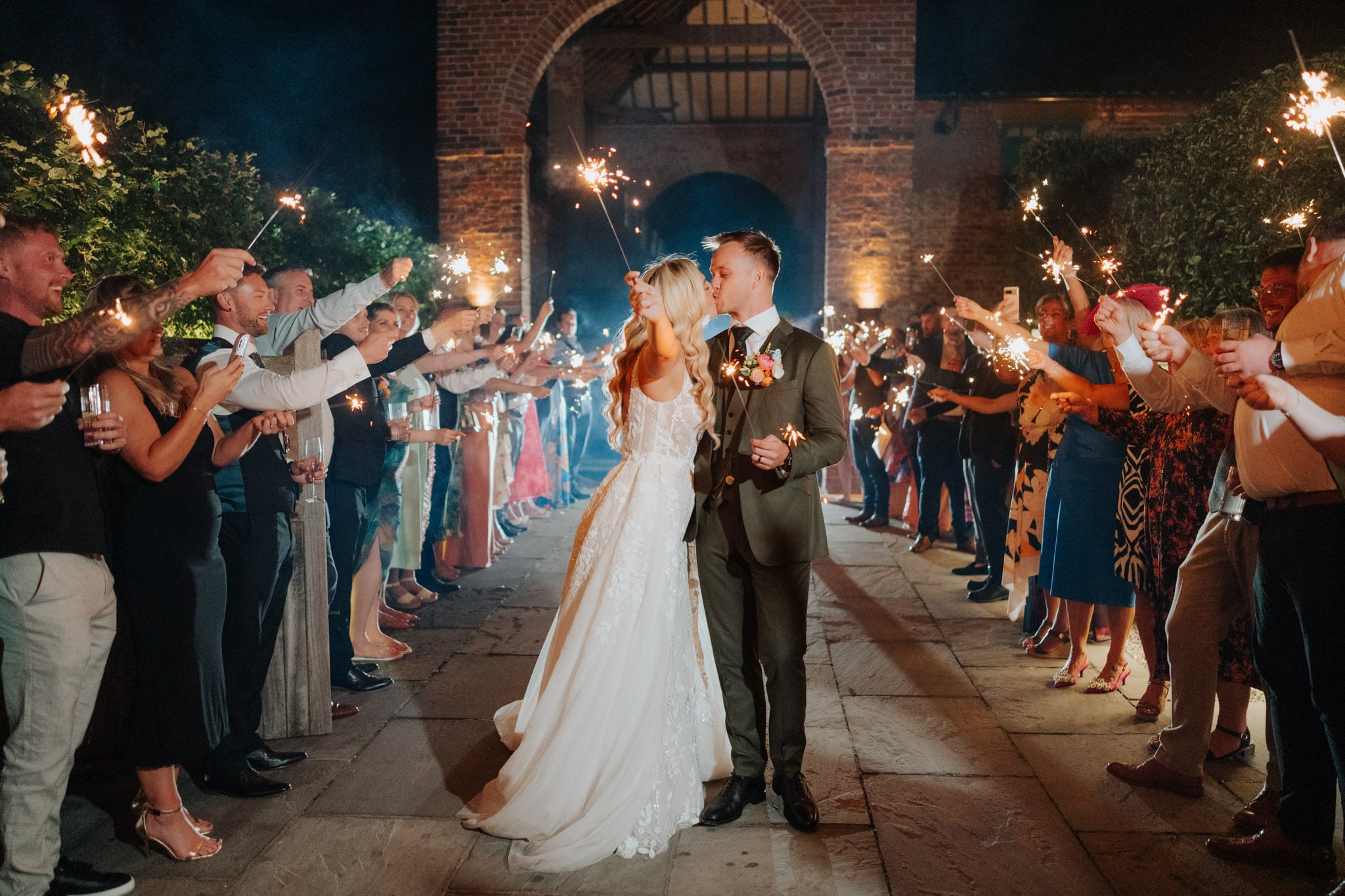 A bride and groom share a kiss outdoors at night, surrounded by guests holding sparklers and cheering. The couple stands in the center, illuminated by warm lights, with a brick archway in the background.