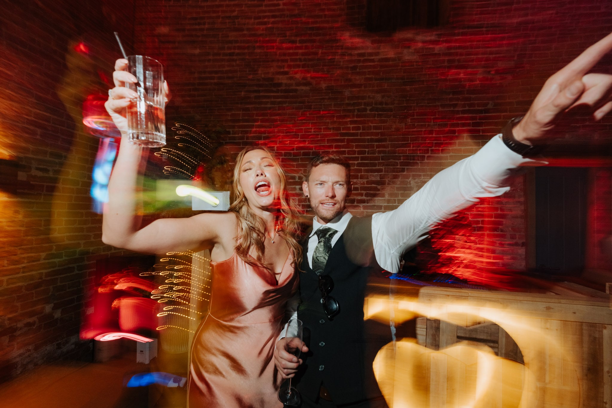 A woman in a satin dress and a man in a suit celebrate at a party, smiling and holding drinks, with colorful lights and a blurred background creating a lively atmosphere.