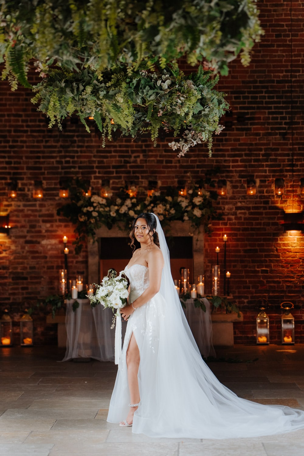 A bride in a white strapless gown with a long veil stands indoors, holding a bouquet. She is posed in front of a decorated brick wall with candles, greenery, and hanging foliage overhead.