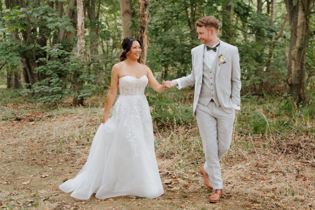 A bride in a strapless white gown and a groom in a light grey suit walk hand in hand in a wooded area, smiling at each other with trees and greenery in the background.