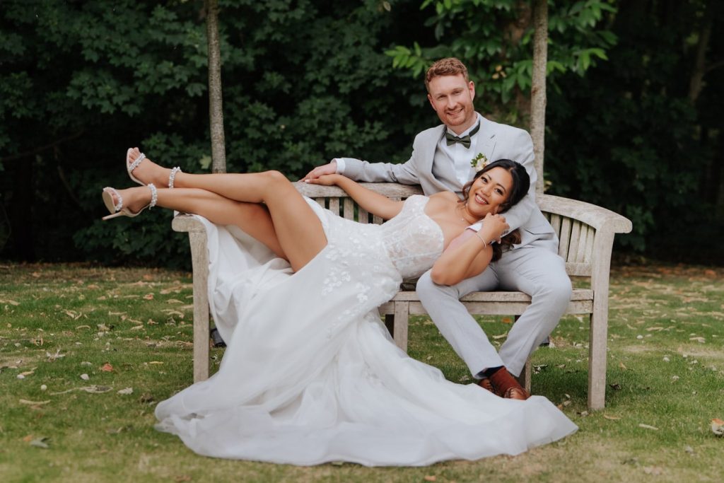 Bride with a beautiful split in her wedding dress lies on the groom whilst both are sat on the bench in the garden at Hazel Gap Barn