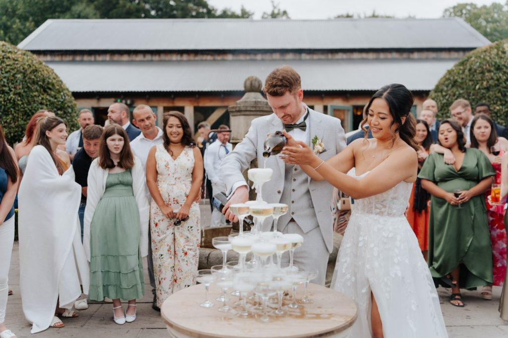A bride and groom pour champagne into a pyramid of glasses during their outdoor wedding celebration, surrounded by smiling guests dressed in formal attire.