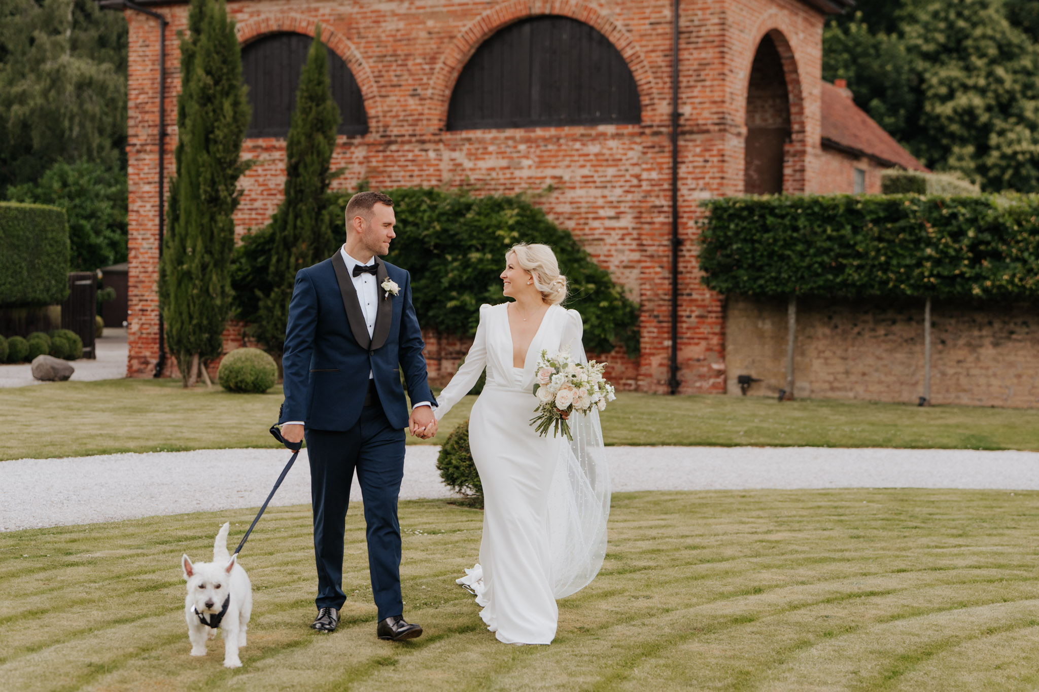 A bride in a white dress and a groom in a navy tuxedo walk hand in hand on grass, smiling at each other, with a white dog on a leash with Hazel Gap Barn behind