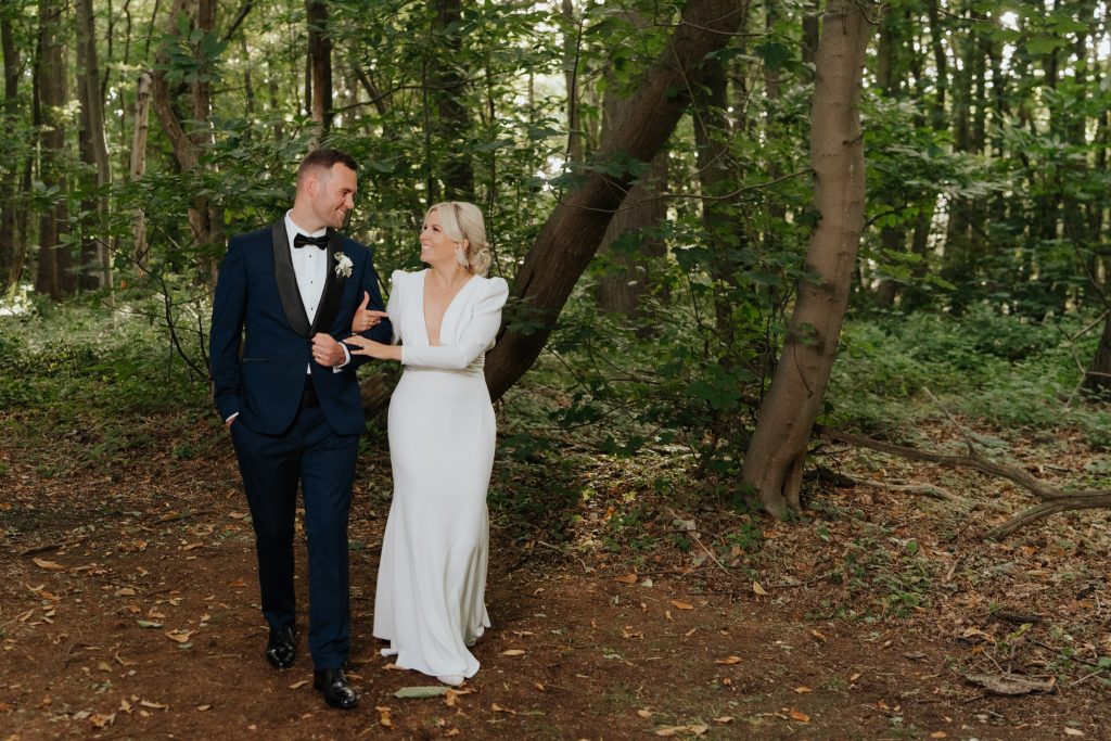 A bride in a long white dress and a groom in a navy suit walk arm in arm, smiling at each other, along a path in a green, sunlit forest.