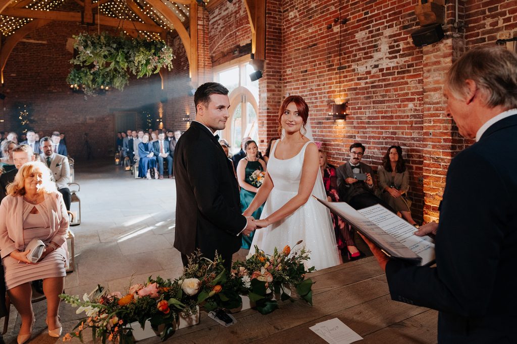 A bride and groom stand hand in hand at the altar in Hazel Gap Barn, a rustic, brick-walled venue, smiling as an officiant reads from a script. Guests watch the ceremony while floral arrangements decorate the table in front.