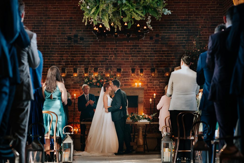 A bride and groom kiss at their Hazel Gap Barn indoor wedding ceremony, surrounded by standing guests. The venue features brick walls, candlelight, and greenery above. A man in a suit stands behind them, clapping by a table with flowers.