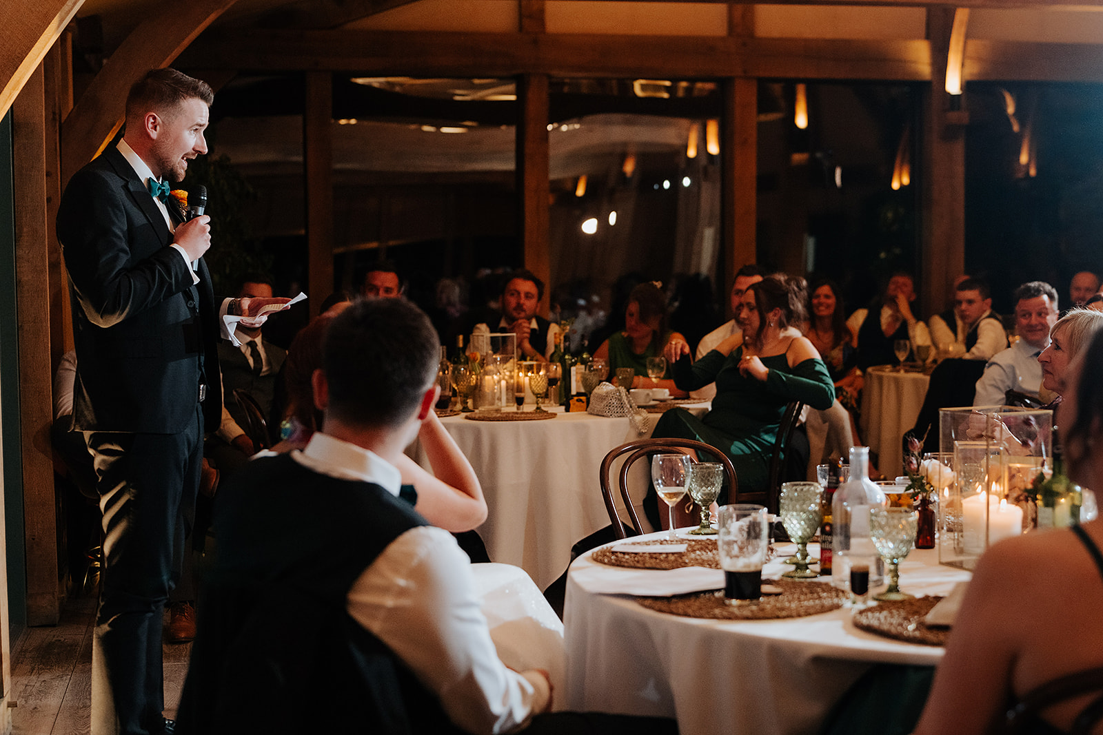 best man stands with a microphone, giving a speech at a warmly lit wedding reception in the Oak Room at Hazel Gap Barn. Guests seated at round tables listen attentively, surrounded by candles and elegant table settings.