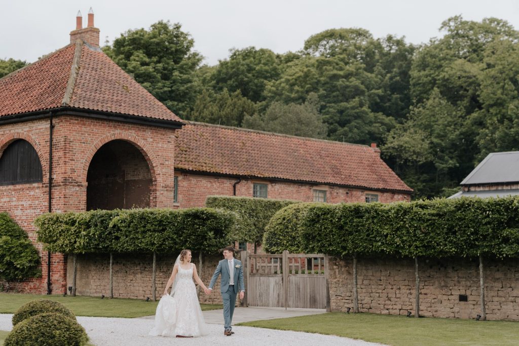 A bride and groom hold hands while walking outside on a stone path. They are outside Hazel Gap Barn and surrounded by neatly trimmed hedges and trees.