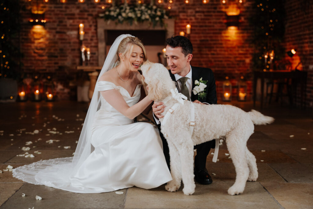 A bride and groom kneel indoors, smiling and petting a large, curly white dog wearing a ribbon. The setting is warm with candles, string lights, and rose petals scattered on the floor.