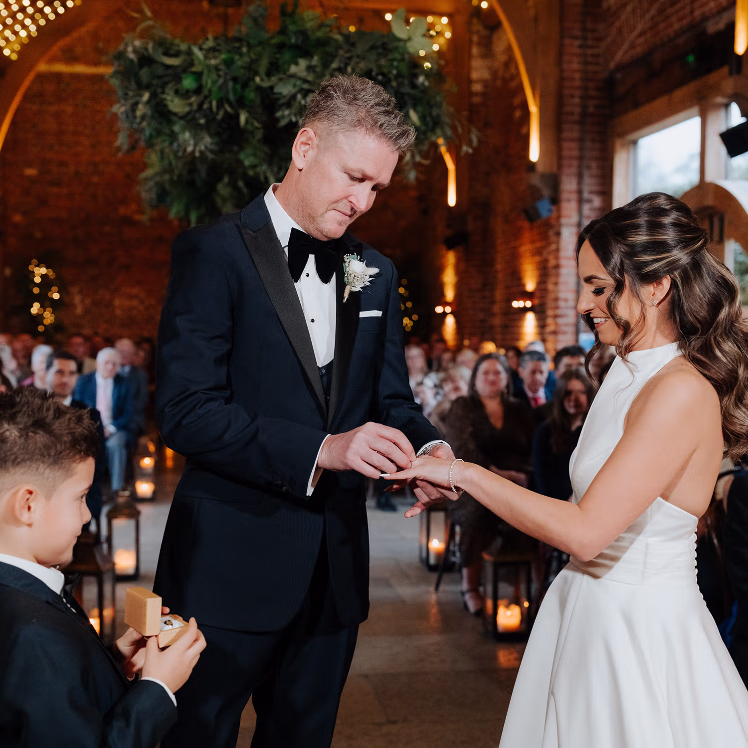 A groom in a black tuxedo places a ring on the brides finger during a wedding ceremony at Hazel Gap with their young son as a ring bearer nearby and guests seated in the background.