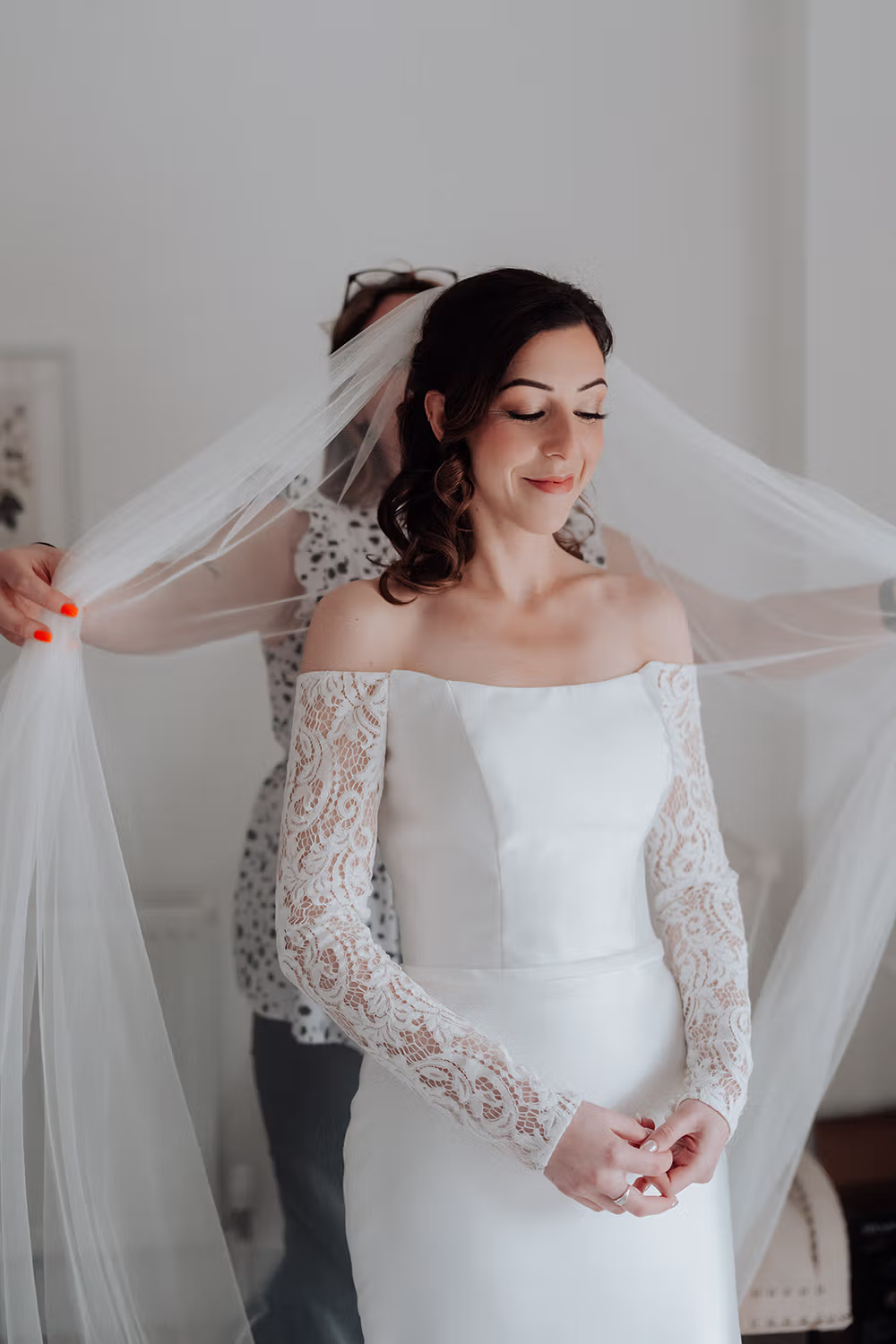 A bride in a white, off-shoulder wedding dress with lace sleeves smiles softly as another person adjusts her veil from behind in a bright, softly lit room.