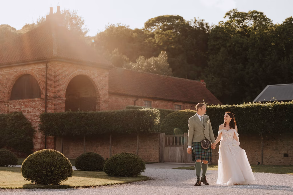 A bride in a white dress and a groom in a green suit with a kilt walk hand in hand outside Hazel Gap Barn, with sunlight streaming through the trees in the background.