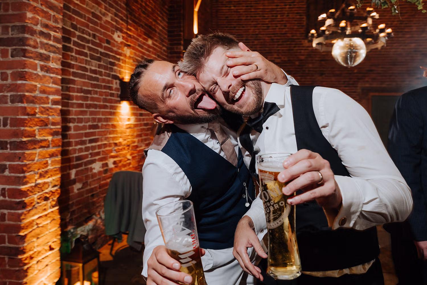Two men in formal wear laugh and pose playfully, holding beers at a lively indoor event with brick walls. One man sticks out his tongue and hugs the other’s head, both appearing joyful and energetic.