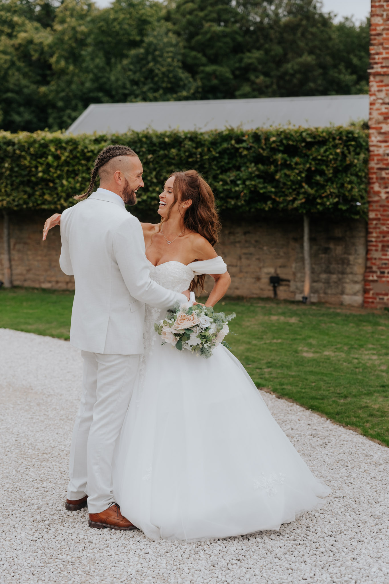A bride and groom stand outside, smiling and gazing at each other. The groom wears a light suit and the bride wears an off-shoulder white gown while holding a bouquet of flowers. Greenery and a brick wall are in the background.