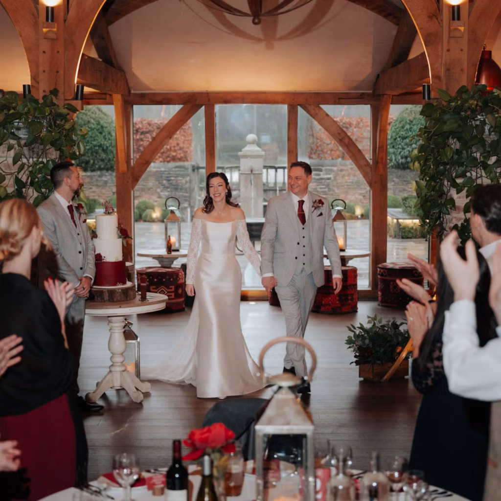A bride and groom, holding hands and smiling, enter a rustic wedding venue with large windows, wooden beams, and guests clapping. A two-tier cake stands on a table beside them. The room is warmly decorated with plants and candles.