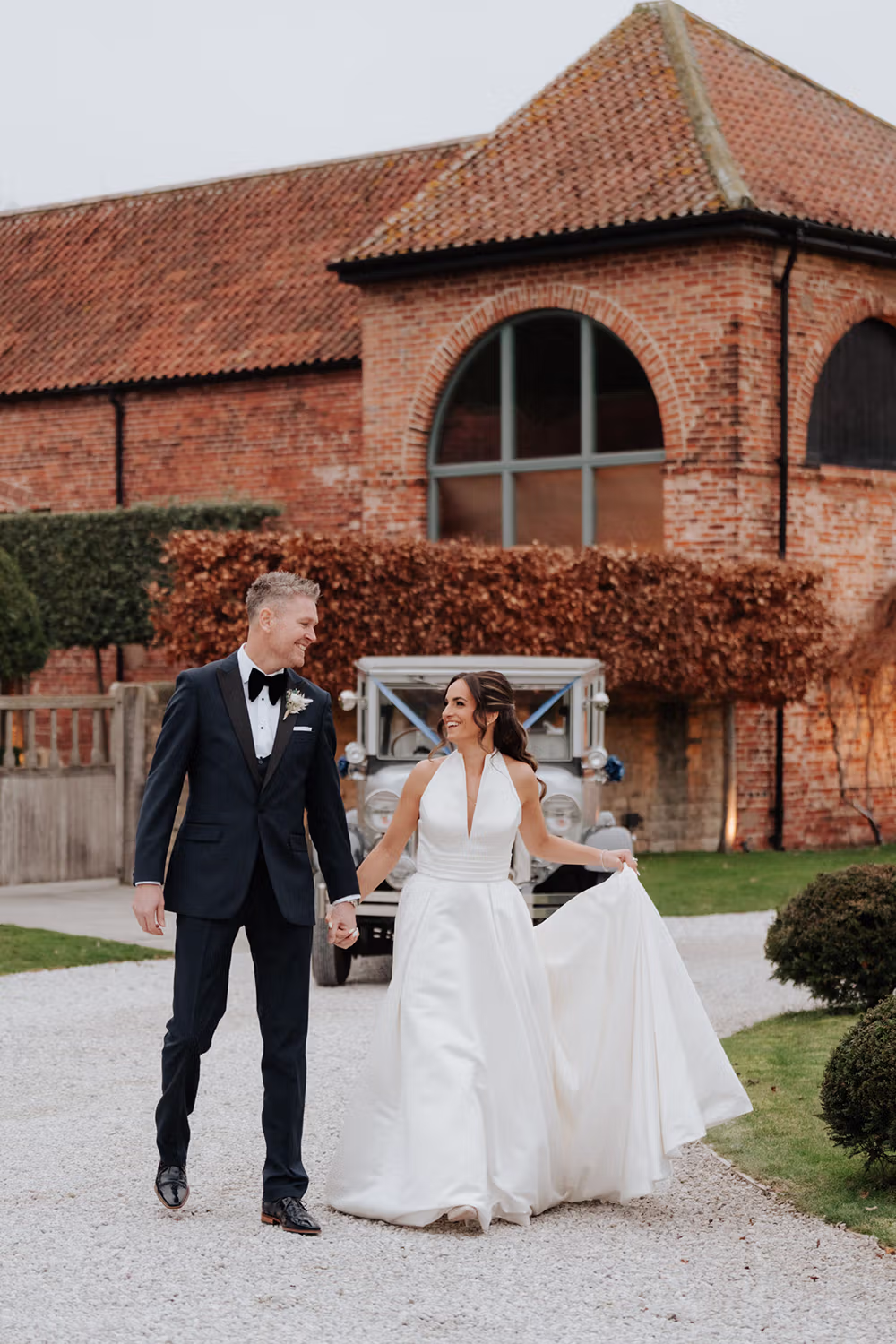 A bride in a white gown and a groom in a black tuxedo walk hand in hand, smiling at each other, outside a rustic brick building with a vintage car in the background.