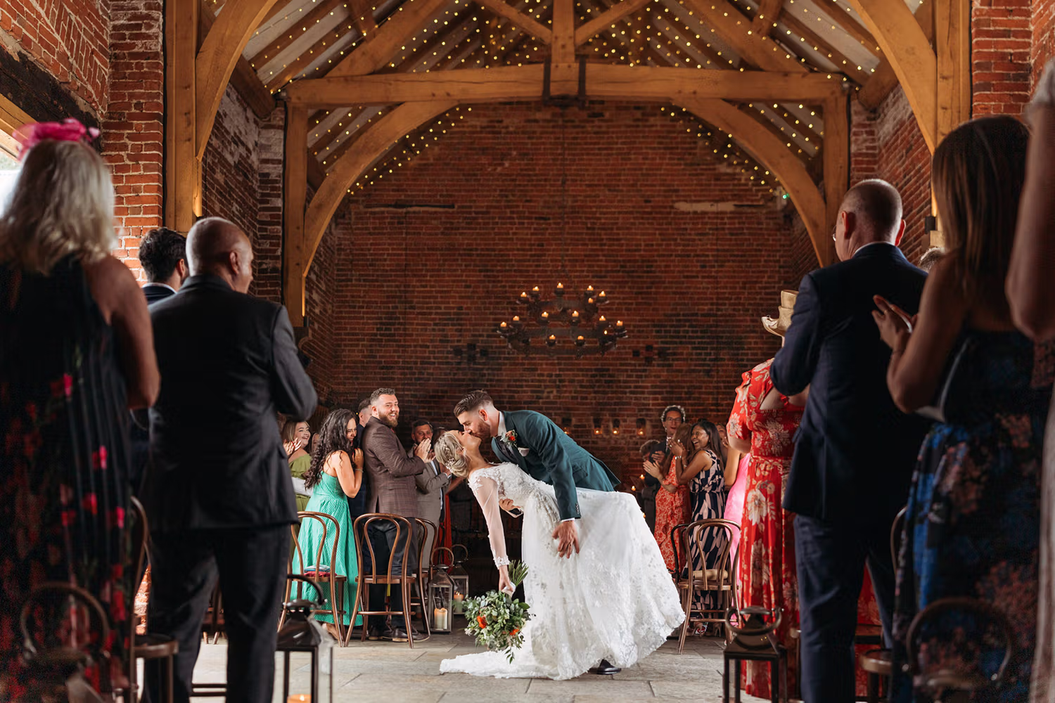 A bride and groom share a dramatic kiss at the end of the aisle inside a rustic barn, surrounded by cheering and applauding guests. Warm lights and wooden beams create a romantic atmosphere.