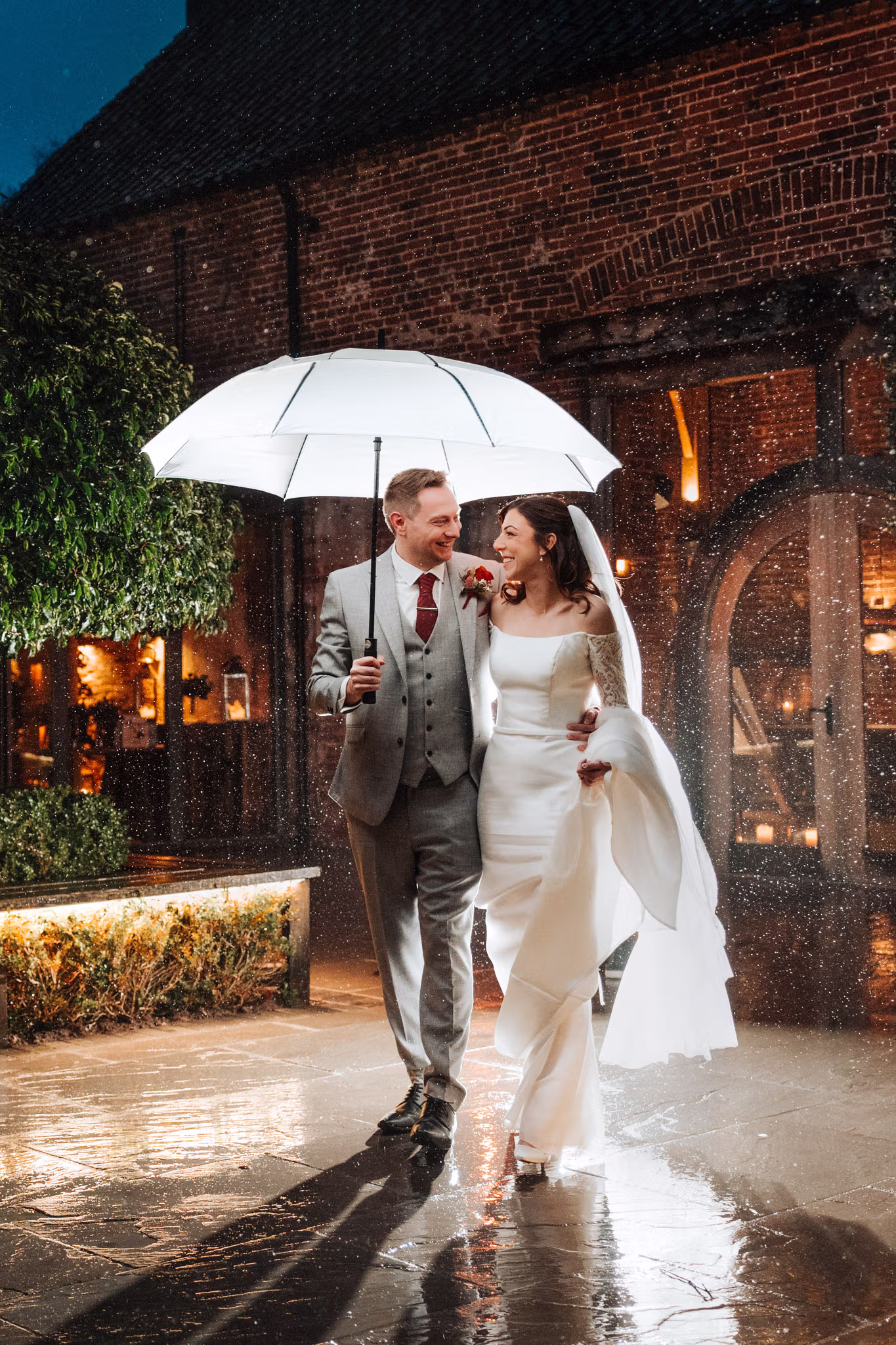 A bride and groom smile while walking under a white umbrella in the rain at night in the courtyard of Hazel Gap Barn