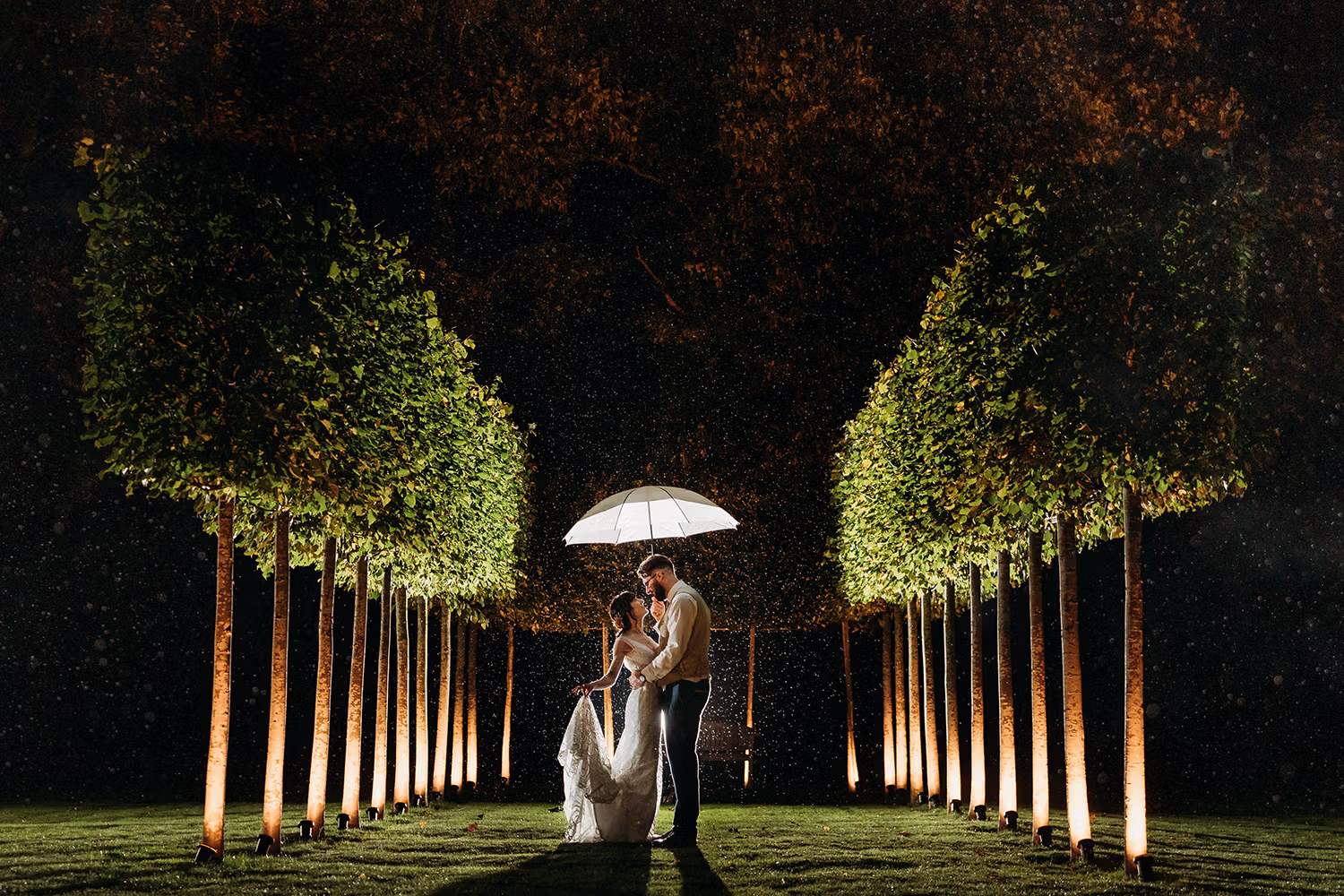 A bride and groom stand under a white umbrella at night, surrounded by rows of illuminated trees in the garden at the back of Hazel Gap, with rain softly falling around them and creating a romantic, magical atmosphere.