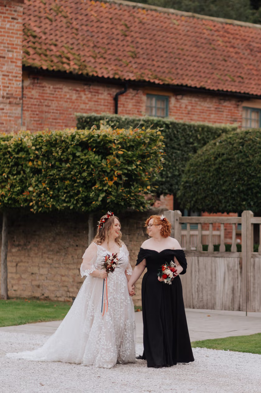 Two brides stand outdoors, holding hands and smiling at each other. One wears a white floral wedding gown and the other wears a black off-shoulder dress; both hold small bouquets and are surrounded by greenery and rustic buildings.
