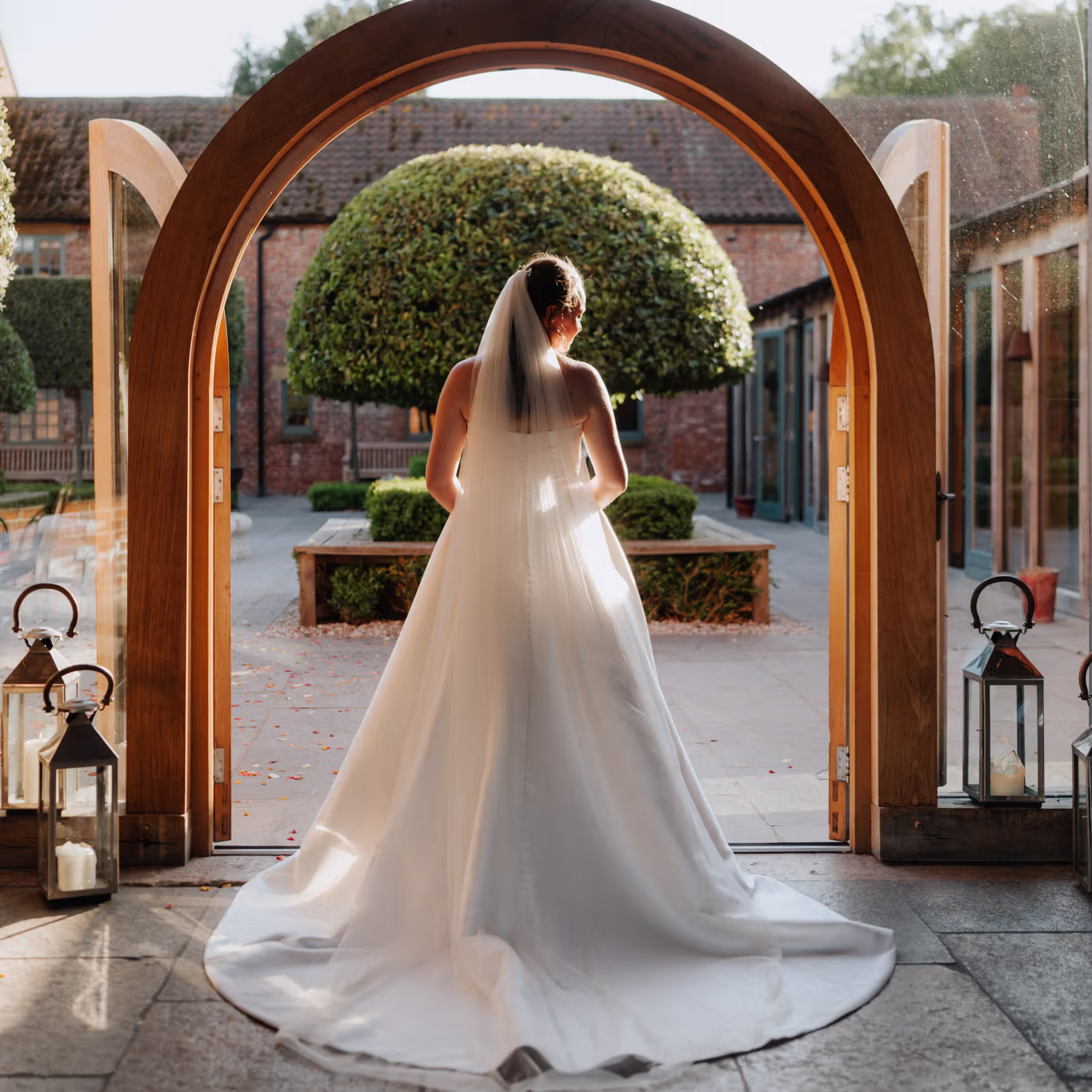 A bride in a white dress and veil stands in a sunlit doorway, facing a courtyard with trimmed trees and brick buildings. Lanterns with candles decorate the entrance.