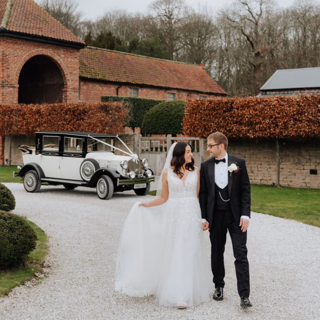 A bride in a white gown and a groom in a black tuxedo hold hands and walk on a gravel path, with a vintage white car and Hazel Gap Barn in the background.