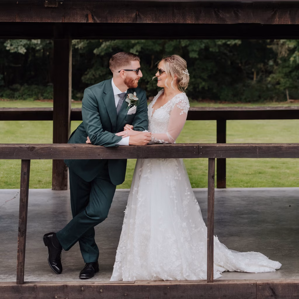 A bride in a white lace gown and a groom in a green suit, both wearing sunglasses, stand smiling and holding hands under a wooden pavilion with green grass and trees in the background.