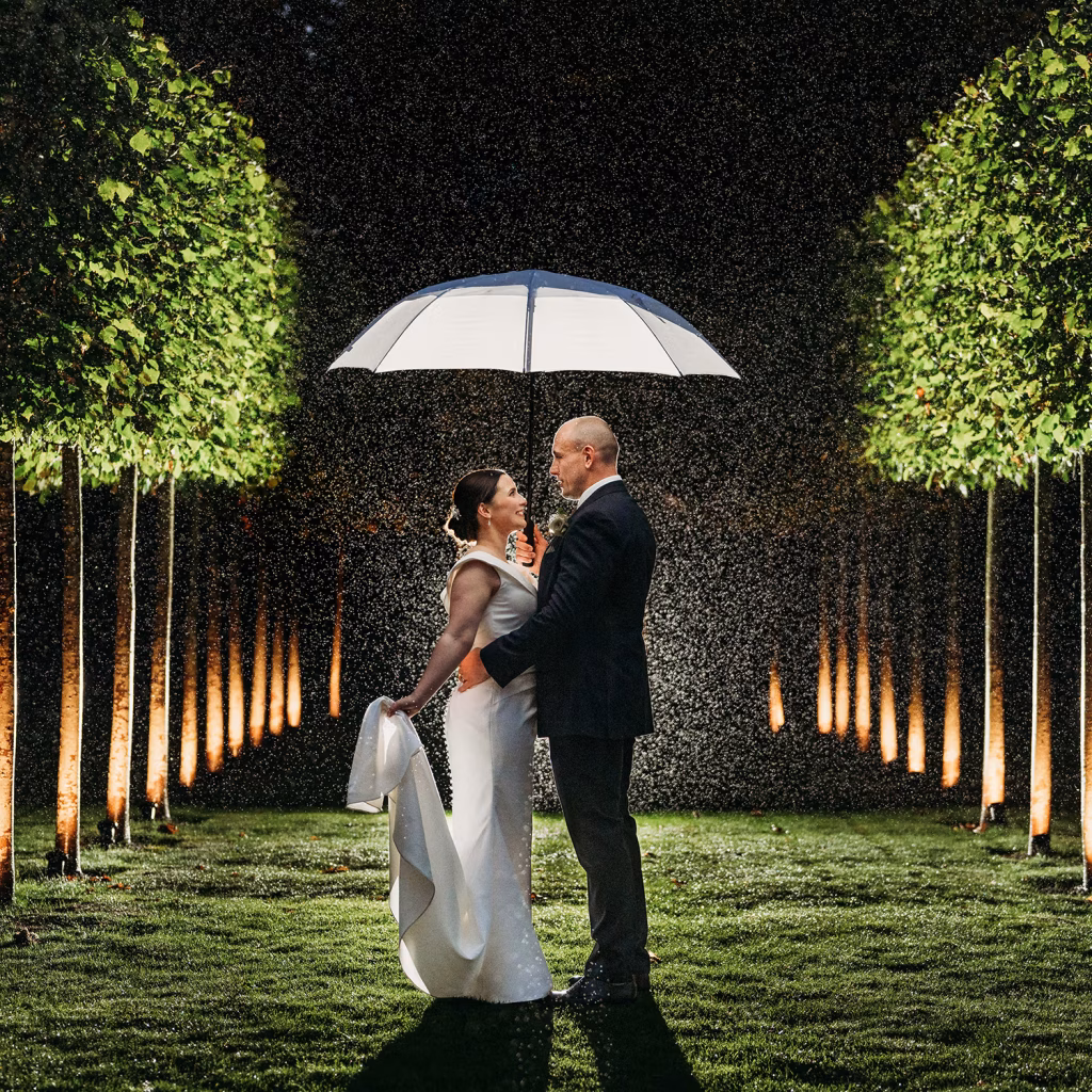 A bride and groom stand close together under a glowing umbrella at night, surrounded by lit trees and falling rain, smiling as they look into each others eyes on a lush green lawn.