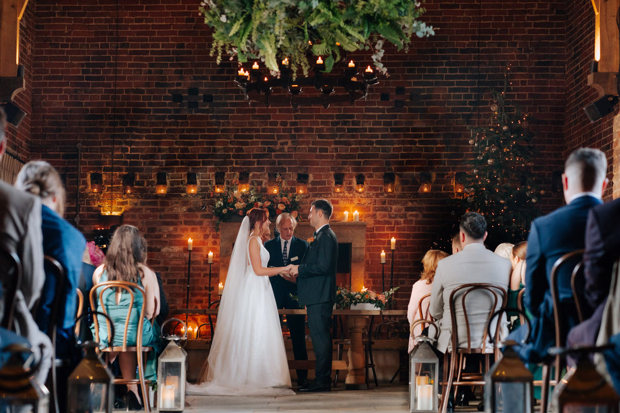 A bride and groom stand at the altar holding hands during a candlelit wedding ceremony in a rustic, brick-walled venue, surrounded by seated guests. Greenery and lights decorate the space.