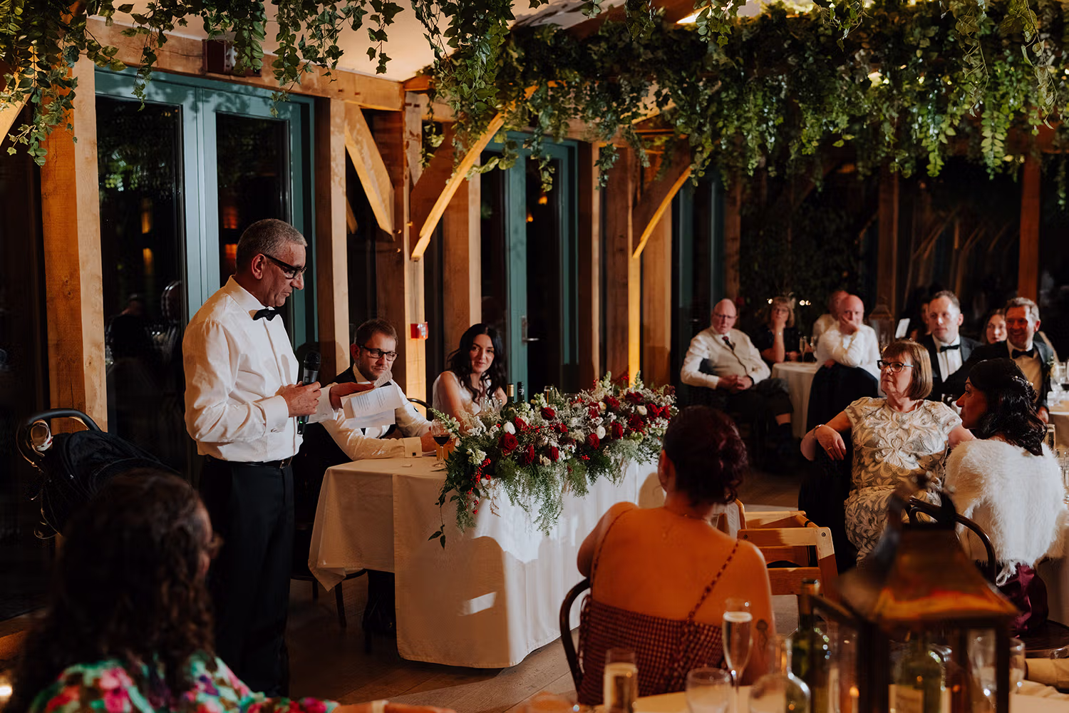 father of bride in a white shirt and bow tie gives a speech at a wedding reception at Hazel Gap Barn, standing beside a table with the bride and groom. Guests seated around listen attentively, under hanging greenery and warm lighting.